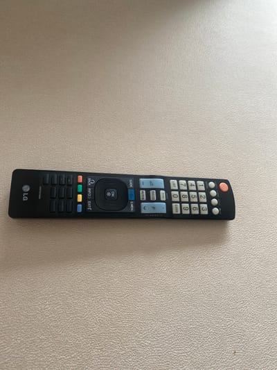 This media file is a photograph of an LG television remote control resting on a textured, light-colored wall. The remote is black with various colored buttons. At the top, there are several circular buttons, including a prominent orange one. Below these are the numerical buttons from 0 to 9, along with "LIST" and "Q.VIEW". Further down, there are blue buttons labeled "RATIO" and "P", and a white "MUTE" button. A central navigation cluster features "GUIDE", "Home", and "Q.MENU" buttons, with a circular OK button surrounded by directional arrows in the middle. Below this are "BACK", "INFO", and "EXIT" buttons, accompanied by four colored rectangular buttons. The remote has a serial number "AKB73275606" printed near the bottom, and the LG logo is visible at the very end. The lighting suggests it is daytime, and the setting is indoors, likely a home or office, given the presence of a remote control. There are no people or animals visible in the image.