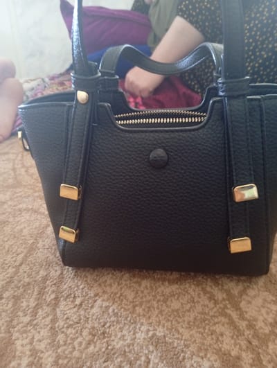 A close-up shot focuses on a black pebbled leather handbag with gold-toned hardware, including zipper teeth, clasps, and decorative elements. The bag is positioned on a textured, carpeted surface. In the blurred background, hints of a person’s legs and arm, along with what appears to be a purple garment, are visible. The scene is indoors. There is no information in the image to suggest a specific location in the city of Hisor, Tajikistan.