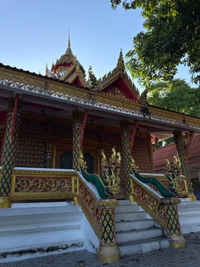 The image depicts the exterior of a Buddhist temple in Baan Tha Po, Thailand. The temple features ornate golden and red architectural details, including decorative rooflines and intricate carvings. Two prominent staircases with balustrades adorned with green serpent-like figures lead up to the temple entrance. The building's facade is decorated with a patterned gold and dark blue wallpaper. A large tree with green leaves partially shades the temple on the right side of the frame. The sky is clear blue. No people are visible in the image.
