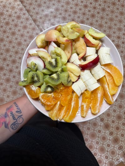 A top-down shot shows a white bowl filled with a colorful fruit salad. The salad contains sliced bananas, orange segments, kiwi slices, and chopped apples or peaches. The fruits are arranged in a visually appealing manner, filling the majority of the bowl. The bowl itself rests on a patterned surface with a beige and brown geometric design. To the right of the bowl, a person's arm wearing a black garment is visible. The person has a tattoo on their forearm with visible text that appears to be in a stylized script, reading "Romiz Vilena". The lighting suggests it might be daytime.