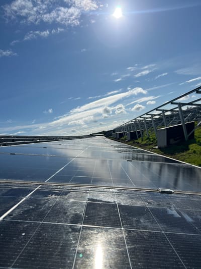 The image depicts a series of solar panels at Grange Ville, Ireland, facing the sky, reflecting clouds and sunlight. The panels appear somewhat dirty. Supporting metal framework is visible. In the background, grass or vegetation is observed.