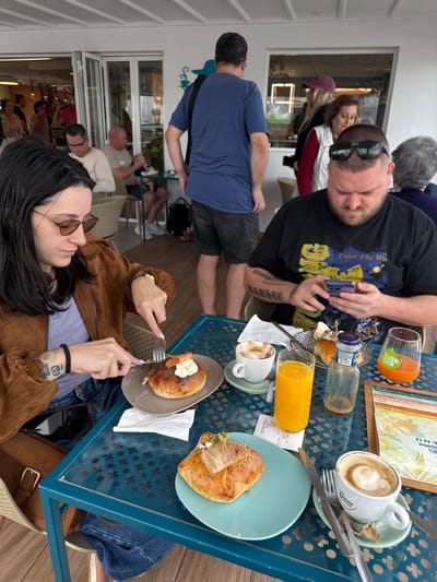 The image depicts a daytime scene at an outdoor cafe or restaurant in Playa Blanca, Spain. In the foreground, two individuals are seated at a teal metal table with a perforated pattern. On the left, a female with dark hair, wearing brown sunglasses, a brown suede-like jacket, and blue jeans, is actively cutting a round pastry with a knife and fork. The pastry, served on an oval brown plate, is topped with white cream, orange fish (likely salmon), and poppy seeds. A white ceramic cup containing a latte or cappuccino and a clear glass of orange juice are also visible on her side of the table. On the right, a male with dark hair, a beard, and dark sunglasses resting on his head, wearing a black t-shirt with a "Enter The Wu" graphic, is looking down at a blue smartphone held in both hands. His table setting includes a light blue plate with a square pastry topped with meat and sesame seeds, a white ceramic cup with a latte/cappuccino, a clear bottle, a glass with a "Fuze Tea" logo containing an orange beverage, and a small black speckled plate with additional pastries. A laminated menu and a receipt are also on the table.
In the mid-ground, a male with his back to the viewer, wearing a blue t-shirt, dark shorts, and a black shoulder bag, stands near a white wall with a blue coffee-themed graphic. To the left, two males are seated at a smaller table. To the right, behind the foreground male, a woman in a maroon hat and another with gray hair are partially visible. The background features white walls, large glass windows or doors, and additional patrons further inside or outdoors, indicating a bright, open cafe environment.