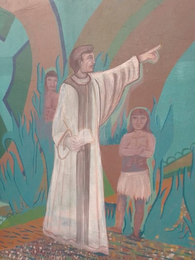The image depicts a mural in Villa Hayes, Paraguay. In the foreground, a light-skinned man wearing a long white robe with a brown central panel gestures with his right arm extended, pointing towards the right. He holds an object in his left hand. To his right stands a Native American girl with arms crossed, wearing a light-colored skirt. Behind and to the left of the man, a second Native American figure is partially obscured. The background is a stylized landscape with blue-green and brown shapes representing plants and land formations. A faint rainbow-like arc appears in the upper right corner. The mural has a textured appearance.