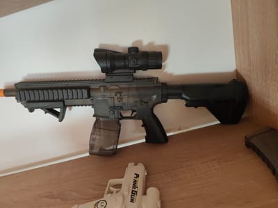 A black, assault-rifle style toy gun with a scope and a transparent magazine is displayed on a shelf. To the right of the toy gun, another black magazine is visible. Below the toy gun, a white toy handgun with the words "Frank Gun" printed on it is also on the shelf. The background consists of a white wall and a wooden surface. The image was taken in Edineț, Moldova.
