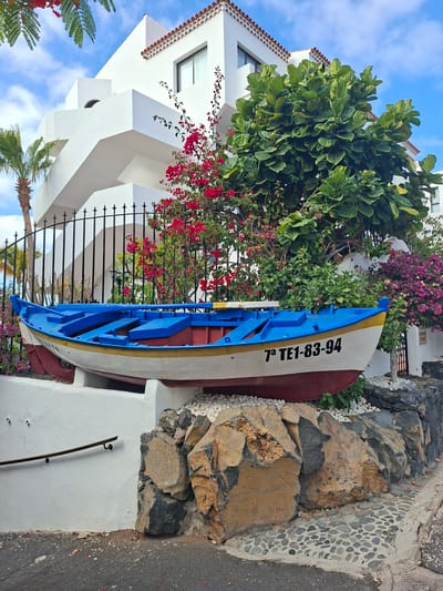 A blue and white fishing boat with red trim is displayed on a stone pedestal in front of a white building with stairs and lush green foliage. The boat has black lettering on its side that reads "7ª TE1-83-94." The building has a traditional Spanish tiled roof. The surrounding environment includes palm trees, flowering bushes in red and purple, and large rocks. The scene is outdoors on a sunny day with a blue sky and scattered clouds. The location is in Santiago del Teide, Spain.