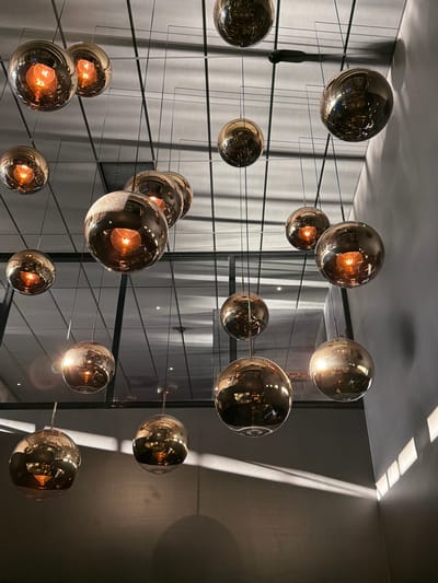 The image shows a close-up, low-angle view of a cluster of spherical, reflective pendant lights hanging from the ceiling in what appears to be a modern interior space. The lights have a metallic, bronze or copper hue and contain visible light bulbs emitting a warm, orange glow. The reflections within the spheres show distorted images of the surrounding environment, including structural elements and other lights. The ceiling is a grid-like structure with dark lines, and there is a light-colored wall to the right with a pattern of sunlight casting shadows. The overall impression is one of contemporary interior design, with a focus on lighting and reflective surfaces. The context of Östersund, Sweden, is not evident from the visual information provided in the image itself.