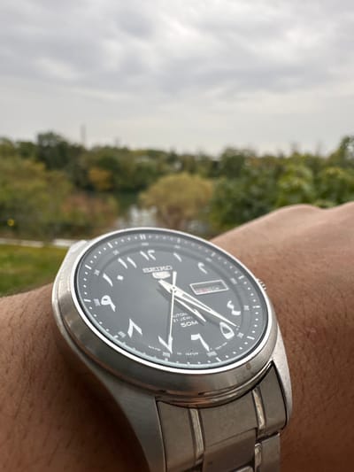The image presents a close-up, angled view of a silver-toned Seiko 5 automatic wristwatch on a human left wrist. The watch features a black dial with white Eastern Arabic numerals and white hour and minute hands, indicating a time of approximately 2:40. A day/date complication displays "SUN" in red. The dial also bears the inscriptions "SEIKO", "AUTOMATIC 21 JEWELS 50M", and "MADE IN JAPAN". The watch's stainless steel case and link bracelet exhibit minor surface abrasions. The background is out of focus, revealing an outdoor setting under an overcast sky. A body of water is partially visible behind a dense line of green and scattered autumnal trees. A faint, slender, dark-colored tower-like structure is discernible in the distant left, rising above the treeline. The scene is located in Épinay-sur-Seine, France.