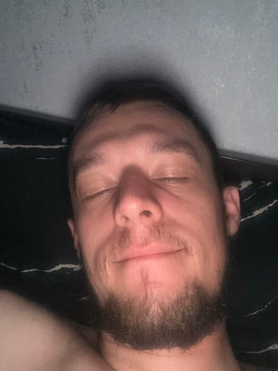 A close-up, overhead, selfie-style photograph features a man with closed eyes and a slight smile. He has short brown hair and a beard. The background consists of a dark fabric with a white wavy pattern and a textured, gray surface. The lighting is warm, illuminating the man's face. No specific location within Zhigulyovsk, Russia, can be determined from the image.