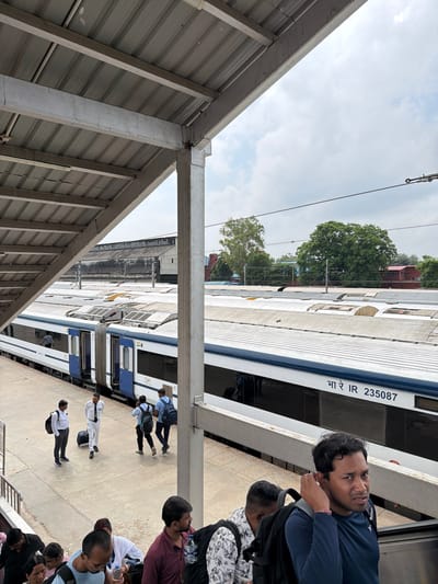 An elevated shot captures a train platform in Lucknow, India, under a partly cloudy sky. A modern blue and white train, identified by the markings "भा रे IR 235078," is parked at the platform. Several individuals are visible on the platform, including people in uniform and passengers with luggage. Some are walking, while others appear to be waiting. A covered walkway with a metal roof extends from the foreground, obscuring part of the sky. In the background, buildings and lush green trees are present. The overall scene depicts a busy railway station environment.