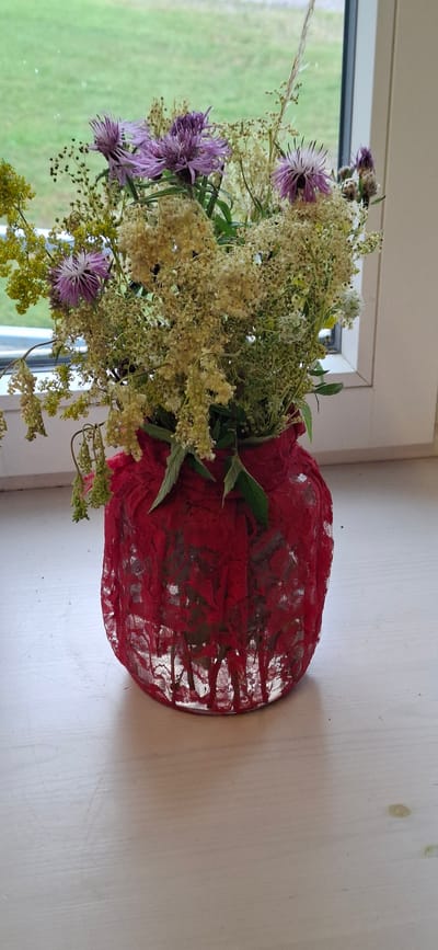 A vertical shot shows a bouquet of wildflowers in a red lace-covered vase placed on a white windowsill. The vase is round and appears to be made of glass or ceramic, with red lace draped over it, creating a textured and decorative effect. The lace has an intricate floral pattern.

The bouquet is composed of various delicate wildflowers. Prominent among them are purple thistle-like flowers with spiky petals, interspersed with clusters of small, airy, pale yellow and white flowers and green foliage. Some wispy grass stalks are also visible, adding to the natural, wild aesthetic of the arrangement. The flowers are arranged loosely, giving them a natural, just-picked appearance.

The background shows a window with a view of green fields under what appears to be natural daylight. The light suggests it is daytime, possibly late morning or afternoon. The setting is indoors, by a window, likely in a home or a rural building in Sääre, Estonia, given the context. The overall mood is tranquil and evokes a sense of nature and simple beauty. There is no visible text in the image.