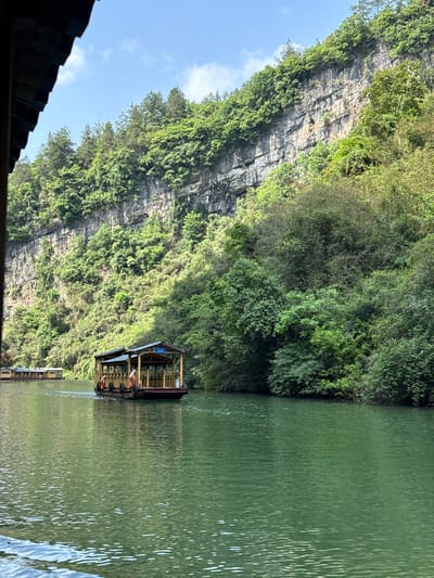 The image shows a river scene in Sanguansi, China. A covered boat is centered in the composition, floating on calm, green water. The right side of the image is dominated by the river, showing slight ripples on the surface. The left side features a large, rocky cliff face covered in lush green vegetation. The top left of the image reveals a sliver of blue sky with a few scattered clouds. Parts of a dark rooftop are visible in the bottom-left corner.