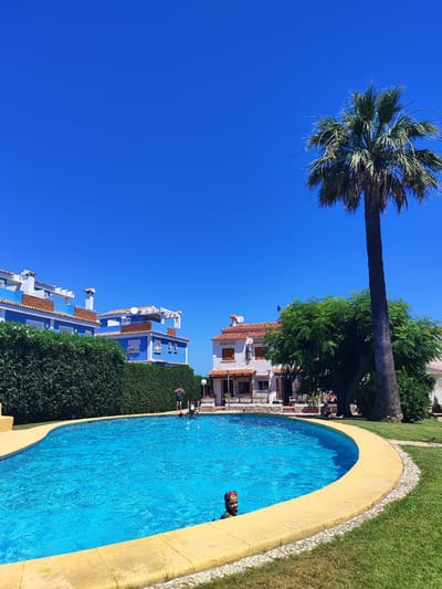 This is a horizontal, eye-level shot of a bright, sunny day in Els Poblets, Spain, featuring a swimming pool and residential buildings. The foreground is dominated by a large, kidney-shaped swimming pool filled with vibrant blue water, its yellow tiled edge contrasting with the lush green grass that surrounds it. A young child wearing goggles is partially submerged in the pool on the left side. On the right side of the pool, near the edge, a person is lying down on the grass, partially shaded by a tall palm tree. In the mid-ground, there's a dense, green hedge separating the pool area from buildings. Beyond the hedge, on the left, are multi-story residential buildings with white roofs and light blue walls, featuring balconies and arched windows. On the right side of the mid-ground, a white building with a terracotta tiled roof is visible, with a tree providing shade in front of it. A tall palm tree stands prominently on the right side of the frame, its fronds reaching towards the clear, bright blue sky. The overall scene conveys a sense of leisure and vacation in a Mediterranean setting.