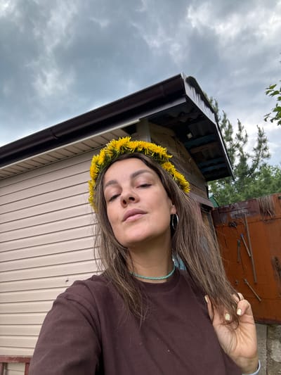 The image shows a woman with a dandelion crown on her head, looking at the camera. She is wearing a dark brown t-shirt and a light blue beaded necklace. Her hair is long and brown, and she is touching a lock of it with her right hand. She has a silver hoop earring in her left ear. In the background, there is a beige-sided building with a dark brown gutter. To the right of the building, there is a wooden fence and some trees. The sky is cloudy and gray. The scene appears to be outdoors, possibly in a rural area or a backyard.