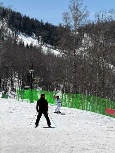 The image depicts a snowy ski slope in what appears to be a mountainous region of Ozernoe, Russia. Two skiers are visible on the slope. One is dressed in black and is actively skiing downhill, while the other is wearing a white ski suit and is standing still. Green safety netting is present on the edge of the slope, near a signpost containing Russian text. Bare trees line the background. A ski lift can be seen running up the mountain in the distance. The sky is clear and blue.