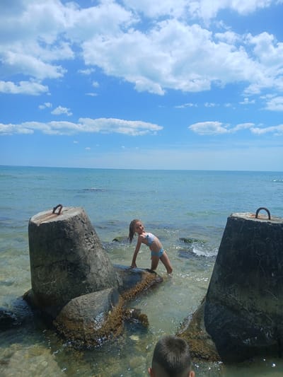 This image depicts a sunny day at the Black Sea coast in Osenovo, Bulgaria. Two individuals are partially visible: a young girl in a blue and white bikini, and the head and shoulders of a young boy on the right side of the frame. The girl is positioned in the water, leaning back with her arms extended, appearing to be playing or posing. The boy is on the shore, with his back to the camera, looking towards the water.

The scene features the clear blue water of the sea, with gentle waves lapping around large, weathered concrete structures that appear to be breakwaters or remnants of an old pier. The sky above is a vibrant blue, dotted with fluffy white clouds. The sunlight creates sparkling reflections on the water's surface and highlights the textures of the concrete. The overall impression is one of a casual summer day spent by the sea. The time of day is likely midday, given the bright sunlight and shadows. No text is visible in the image.