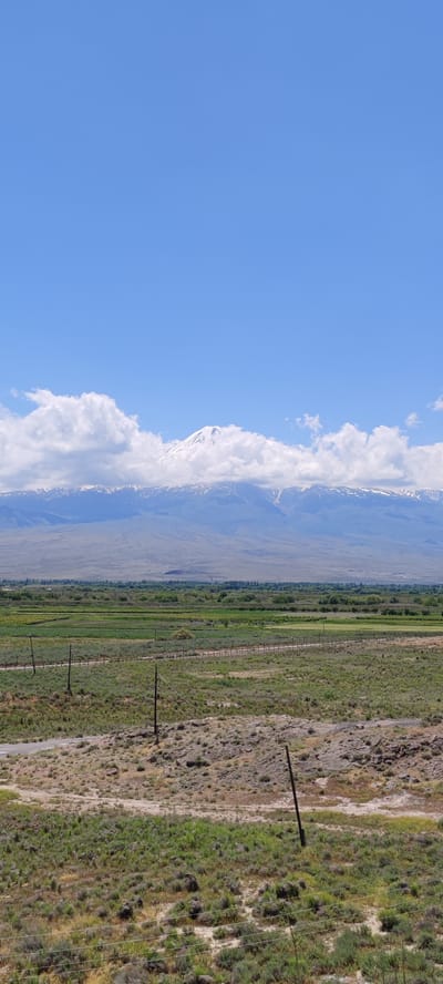 The image shows a panoramic view of the landscape in Lusarat, Armenia. The upper portion of the image is dominated by a clear blue sky with scattered white clouds. Below the sky, a majestic snow-capped mountain range stretches across the horizon, with Mount Ararat appearing to be the most prominent peak. The mountains are partially obscured by fluffy white clouds.

The middle ground of the image features a vast expanse of agricultural fields, with a mix of green crops and lighter-colored fields, suggesting different stages of growth or types of cultivation. A fence line runs horizontally across the fields, and several utility poles are visible, standing amidst the vegetation.

The foreground of the image consists of dry, rocky terrain covered with sparse green and brown vegetation, including bushes and grasses. More utility poles are scattered across this area, and a dirt road or path can be seen winding through the landscape. The overall scene depicts a rural and natural environment with agricultural activity in the mid-distance and a prominent mountain backdrop.