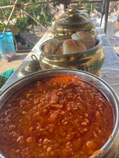 The image shows a food stall in Mira-Bhayander, India, likely set up outdoors. The focus is on a metal container filled with a thick, orange-red gravy, possibly a spicy Indian curry or bhaji. Adjacent to it are two brass containers with lids, one open to reveal several soft, white buns inside. The setup suggests a food offering, possibly Pav Bhaji. In the background, there is a glimpse of the environment including a table, foliage, and a partially visible blue container or cage. The scene appears to be during the day, with natural light illuminating the food and the surroundings.