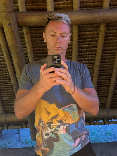 A male individual, appearing Caucasian with short, bleached blonde hair, is depicted taking a selfie within a structure in Sakti, Indonesia. He holds a dark-colored smartphone, featuring a multi-lens camera array and a black pop-socket, in both hands directly in front of his torso. His expression is neutral. He wears a dark grey short-sleeved t-shirt with a complex, multi-colored graphic print featuring surreal elements, including large yellow forms, a figure with a panda head, and abstract shapes. A braided red-brown bracelet is visible on his left wrist. The immediate background shows internal architectural elements constructed from bamboo, including diagonal support beams and a ceiling composed of woven thatch material. Below the structure's frame, a light blue-grey ground or water surface and indistinct green foliage are visible.