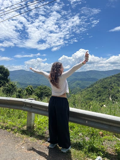 A young woman with curly brown hair is standing on the side of a road, facing away from the camera, with her arms outstretched and raised towards a bright blue sky filled with white, fluffy clouds. She is wearing a white long-sleeved top, black loose-fitting pants, and light blue athletic shoes. In her right hand, she holds a colorful object, possibly a phone or a small device. The road is paved with asphalt and bordered by a metal guardrail. Lush green vegetation, including trees and grasses, grows on the embankment behind the guardrail. In the background, rolling hills covered in dense green forest stretch as far as the eye can see under the expansive sky. The scene suggests a location with natural beauty and an open, elevated viewpoint, consistent with mountainous terrain. This scene is located in the city of Son Dien, Vietnam.