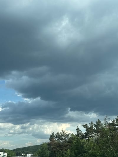 The image shows a view of the sky from a highway, with dark storm clouds dominating the upper portion. Below the clouds, there are patches of blue sky and lighter, more scattered clouds. The horizon is lined with green trees and a rolling hillside. On the highway, several white trucks and cars are visible, along with some road signs. The overall impression is of an overcast or approaching storm.