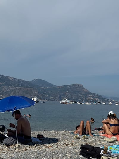 A wide shot captures a pebble beach in Saint-Jean-Cap-Ferrat, France, on a sunny day with a hazy sky. The water is calm and reflects the overcast conditions. In the foreground, a blue umbrella provides shade for a man sitting on a towel. He is wearing black shorts and appears to be looking at his phone. Nearby, a black helmet and a grey blanket are visible. Further down the beach, two people are partially submerged in the water. To the right, a couple is relaxing on towels. The woman on the right is wearing a blue bikini and a white cap, while the man next to her is lying on his back with his legs raised. Several boats and yachts are anchored in the bay, with a mountainous coastline in the background dotted with buildings. The overall scene depicts a leisurely day at the beach.