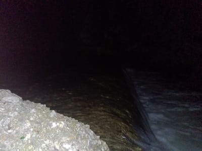 This media file presents a dark, low-light scene, likely captured at night, depicting a body of water flowing next to a rocky bank. In the bottom left, a rugged, light-colored bank made of rocks or concrete is prominent, displaying varied textures and shades of gray and white, with a small green leaf visible among the rocks. The water, which appears dark, occupies the central and right portions of the image. Subtle patterns on the water's surface suggest movement and current. Towards the right, the water flows over a sharp, linear edge, creating a small waterfall or weir, where the water appears frothy and lighter in color as it cascades downwards. The upper parts of the image are extremely dark, providing minimal detail and emphasizing the low-light conditions. No people or visible text are present. The setting is an outdoor environment in Tahlequah, United States of America, likely featuring a river, stream, or other water channel.