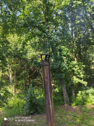 The image is a medium shot of a metal chimney pipe standing vertically in front of a dense forest. The pipe is reddish-brown and appears to be made of copper or a similar metal, with a dark cap on top. It is positioned in the foreground, slightly off-center to the right. Behind the pipe, a lush green forest fills the frame, with sunlight filtering through the leaves. The trees are varied, with some appearing to be deciduous and others possibly coniferous. The ground in the foreground is covered with grass and fallen leaves, and there is a hint of a path or disturbed earth. The overall impression is one of a natural, outdoor setting, likely in a rural or semi-rural area. The image also contains a watermark at the bottom left corner that reads "SHOT ON MI 9T AI TRIPLE CAMERA". There are no people visible in the image. The scene appears to be in Blokhino, Russia, a village located in the Ryazan Oblast.