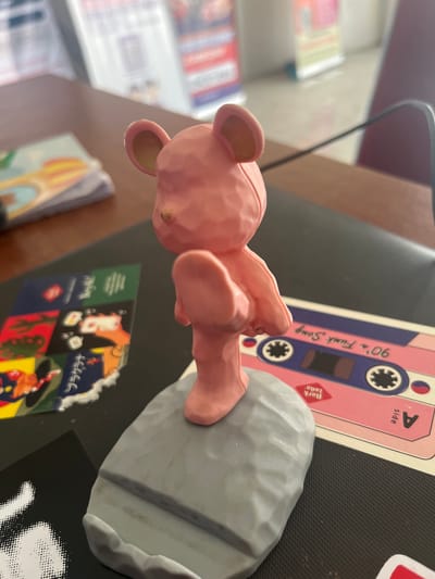 A pink plastic teddy bear figurine is positioned on its side, with its head resting on a grey, rock-like object. The bear is facing left, with its arms and legs extended. In the background, a pink cassette tape with blue accents is visible, featuring text that reads "90's Funk Song" and "A side". Further to the left, out-of-focus stacks of colorful items, possibly books or magazines, are present. The scene appears to be indoors, with a dark, textured surface underneath the objects, which could be a desk or a mat. The lighting suggests it is daytime.