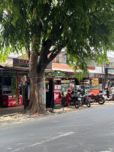 In Siron, Indonesia, a street scene unfolds under the shade of a large tree. Several motorcycles are parked along the roadside in front of a row of shops. One motorcycle, a red sportbike, has a rider wearing a red jacket and helmet. Another motorcycle, a white scooter, is parked further down. A red gasoline pump is positioned near the parked motorcycles. The shops behind the motorcycles appear to be selling various goods, with some displaying signs with Indonesian text. One sign reads "RAKA ANEKA MAKMUR." Bags of snacks hang from a display in front of one of the shops, and a red refrigerator with the brand "Glicoo" is visible. The environment is a bustling street with buildings, a tree, and parked vehicles, suggesting a commercial area.