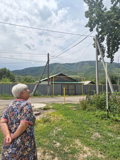 A woman stands outdoors, facing away from the camera and looking upwards at a cloudy sky. She has white hair and wears a floral-print dress with a pink trim. The background features power lines, a metal fence, a building with a green roof, and rolling green hills under a cloudy sky. This scene is located in the city of Tekeli, Kazakhstan.
