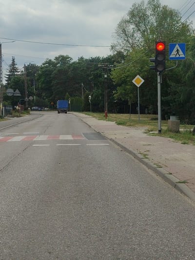 This is an eye-level, medium shot capturing a street scene in Cząstków Mazowiecki, Poland. The foreground is dominated by an asphalt road with a pedestrian crossing marked by red and white stripes. The road curves slightly to the left as it recedes into the distance. On the right side of the road, a paved sidewalk is visible, bordered by grass and trees. A traffic light with a red illuminated signal and a pedestrian crossing sign are positioned on a pole next to the sidewalk. A yellow diamond-shaped warning sign is also present on a separate pole. In the background, a blue truck is driving away from the camera, and a dense line of trees forms the backdrop. The sky is overcast.