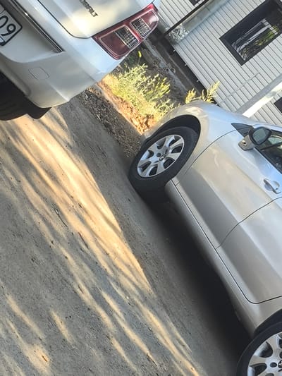The image shows a close-up, angled view of two cars parked on a paved surface. The car in the foreground is a silver sedan, and its front passenger side wheel and door are visible. The car in the background is a white vehicle, and its rear bumper and taillight are partially visible. The scene appears to be outdoors, possibly a street or driveway, with dry vegetation and a building with horizontal siding visible in the background. The lighting suggests it is daytime, with shadows cast across the ground, indicating a sunny day. There is no activity taking place; the cars are parked.
Visible text includes the number "09" on a license plate on the white car and the word "COBALT" on the rear of the white car, which is likely the make or model. The setting appears to be residential, given the building in the background. The overall impression is a still, outdoor scene focused on the vehicles. The location context is Abay, Kazakhstan.