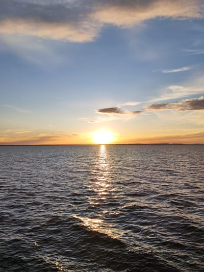 The image depicts a sunset over a body of water. The sun is setting on the horizon, reflecting light onto the rippling water surface. The sky is a mix of blue, orange, and white clouds, with the brightest light emanating from the sun itself. The horizon line is visible in the middle ground, with what appears to be land or a distant shoreline. The image doesn't contain any people. The location is not definitively discernible as Gorushka, Russia, without further context.