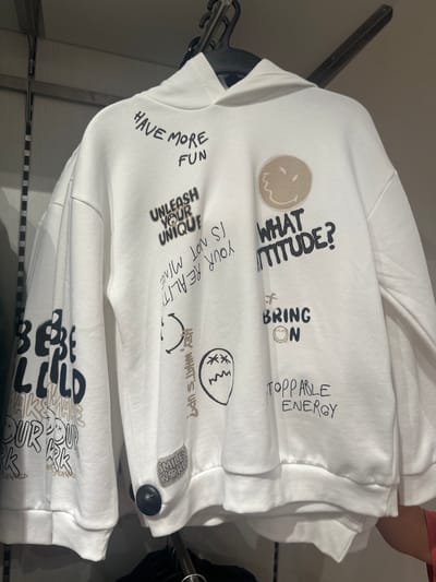 The image shows a white hoodie with various graphic prints and text, displayed on a hanger in what appears to be a clothing store. The prints include phrases like "HAVE MORE FUN," "UNLEASH YOUR UNIQUE," "WHAT ATTITUDE?", "BRING ON," and "STOPPABLE ENERGY." There are also cartoonish drawings of smiley faces and other abstract shapes. On the left sleeve, the text "BE LILD" is visible, along with smaller repeated text. On the lower right side of the hoodie, a small patch with a smiley face and the text "SMILEY WORLD" is attached. The hoodie is made of a textured fabric, and the prints are in black and brown ink. The background is blurred, showing a store interior with hanging racks. No people or specific location details within Polígono Industrial De Quart De Poblet, Spain, are discernable from the image.