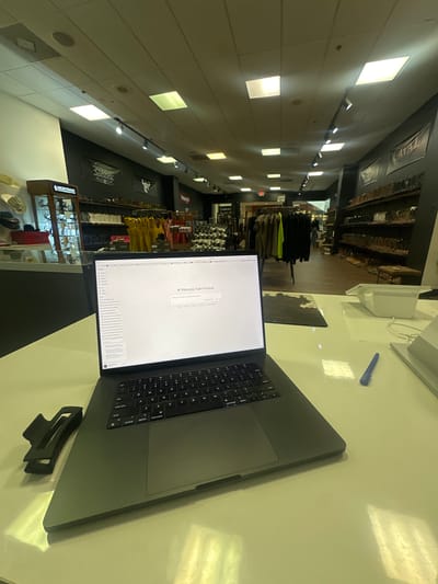 A laptop computer is placed on a white counter in what appears to be a retail store. The screen of the laptop displays a digital document. In the background, shelves are stocked with boots and clothing, suggesting the store specializes in Western wear. The store is well-lit with fluorescent ceiling lights. A blue pen and a black hair clip are also on the counter. The environment is clean and organized.
