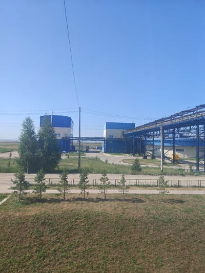 The image shows an industrial facility with several large buildings and extensive pipe racks under a clear blue sky. The buildings are primarily blue and beige. In the foreground, there is a grassy embankment with several young pine trees planted in a line behind a yellow fence. A paved road or pathway runs behind the fence, and beyond that, more industrial structures and vegetation are visible. The overall scene suggests a working industrial site in a rural or semi-rural setting. There are no people or animals visible in the image. The location is stated to be Дүңгілағаш, Kazakhstan.
