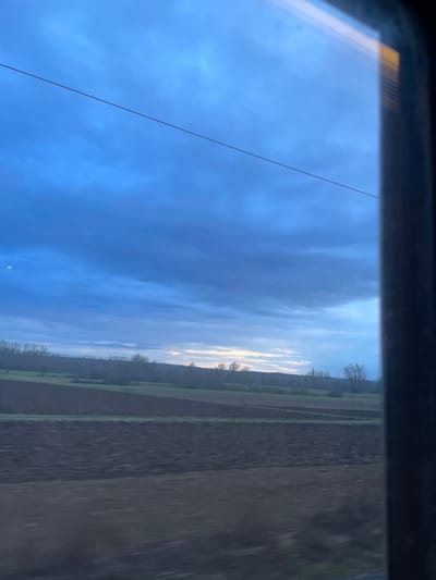 The image, taken from the interior of a vehicle, captures a rural landscape through a window, possibly on a train or car. The perspective suggests motion, with a slightly blurred foreground. The scene depicts a vast expanse of open country under a dynamic sky.

The upper portion of the frame is dominated by a blue sky, ranging from a deep, intense blue in the foreground to lighter, brighter hues near the horizon. Patches of white and grey clouds are scattered across the sky, with some areas along the horizon appearing bright and illuminated, suggesting either early morning or late afternoon sunlight breaking through the cloud cover. A thin, dark wire or cable extends diagonally across the sky from the upper left to the lower right.

The horizon is marked by a distant line of trees, most of which appear bare, indicating either winter or early spring. Beyond these trees, the land stretches out into a mosaic of agricultural fields. Some fields are a rich dark brown, likely freshly ploughed earth, while others exhibit lighter shades of brown and green, possibly indicating different crops or fallow land. The distinct patterns of the fields suggest organized cultivation. The dark, slightly reflective frame of the window is visible along the top and bottom edges of the image.

The overall setting is tranquil and expansive, characteristic of agricultural regions. Given the context of Hochfelden, France, this is likely a view of the French countryside. There is no visible text in the image.