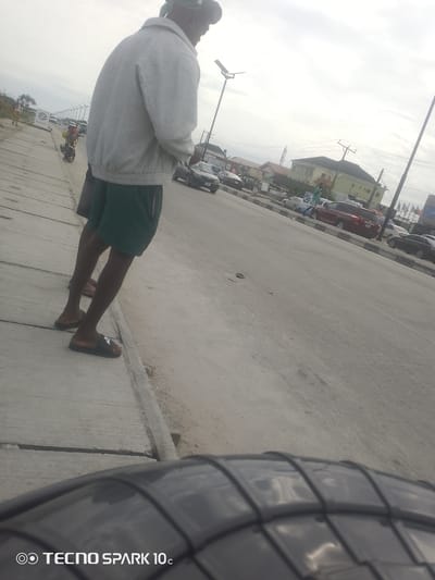 The media file shows a street scene in Awoyaya, Nigeria. The foreground is dominated by the blurred tire of a vehicle, with the "TECNO SPARK 10c" watermark visible in the bottom left corner.
In the middle ground, two people are standing on a concrete sidewalk next to a road. One person is wearing a grey jacket and dark green shorts, with their back to the camera, and the other person is partially visible behind them. The road is paved and has several cars parked along its side, including a red SUV and a black sedan. In the distance, buildings and more cars are visible.
Further in the background, a person is walking on the sidewalk, and a motorcycle is passing by. The scene appears to be set during the daytime, under an overcast sky. The overall impression is a typical street view in a Nigerian town, with vehicles, pedestrians, and residential or commercial buildings lining the road. There are no explicit indications of a specific event or strong emotions.