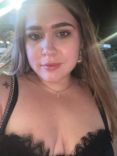 The media file is a close-up, selfie-style shot of a young woman with blonde hair. She is wearing a black lace top and a silver chain necklace with a small cross pendant. Her makeup is natural, with a focus on her eyes and lips. The background is dark and blurry, suggesting an evening setting, possibly outdoors with some ambient lighting. The overall mood appears relaxed and candid. There is no visible text in the image.