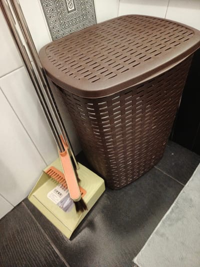 A brown woven plastic laundry hamper with a slatted lid sits on a dark tiled floor next to a light green dustpan and brush set. The broom has a metallic handle and a peach-colored grip. Behind the hamper, a wall is tiled with white squares, and a decorative black and white patterned tile is visible. The scene is indoors and appears to be in a bathroom or laundry area. There is no specific information available about the location being in Usolye-Sibirskoye, Russia, based solely on the visual content of the image.