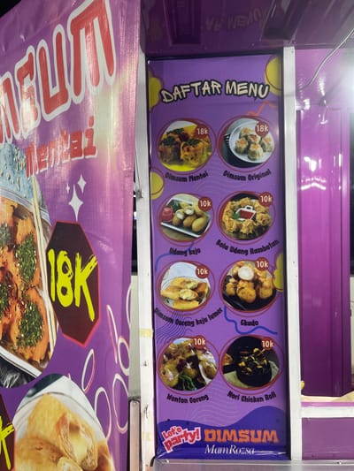 This image shows a food stall that sells dim sum in Anggaswangi, Indonesia. The stall has a purple banner with "MELIMPAH" written in large red letters and "Mentai" in smaller red letters below it. The stall's menu is displayed on a purple board with images of different dim sum dishes and their prices. The prices listed are 18K and 10K, likely referring to Indonesian Rupiah. The menu items include "Dimsum Mentai," "Dimsum Original," "Udang Keju," "Bola Udang Rambutan," "Dimsum Goreng Keju Lumet," "Ekado," "Nonton Goreng," and "Nori Chicken Roll." The stall is decorated with yellow circles and sparkling stars. A portion of a purple food cart is visible on the right side of the image, and a white railing separates it from the food stall. The background is dark, suggesting it might be evening or nighttime.