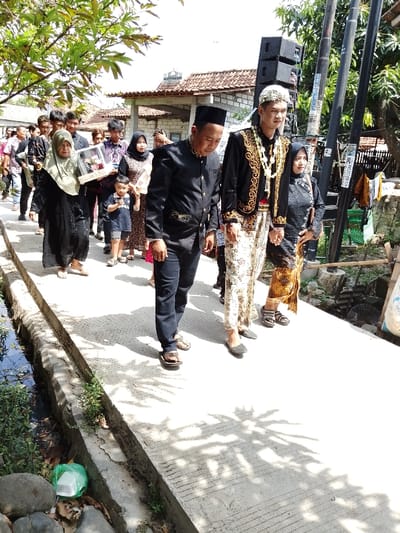 The image depicts a daytime procession of approximately 15 individuals walking on a concrete pathway in Juwana, Indonesia. In the foreground, a man in a black traditional suit adorned with gold embroidery and a black peci (cap) walks forward, looking down, wearing brown flip-flops. To his right, a younger man, identified as a groom, wears a black velvet jacket with extensive gold embroidery, a white patterned sarong, a white patterned peci, and a prominent white floral garland around his neck. He is holding hands with a woman to his immediate right, who wears a dark patterned top and a brown patterned sarong, with dark sandals.
To the left of the leading man, an elderly woman in a long black dress and a light green hijab carries a rectangular, clear-covered box, possibly a gift or dowry, with a brown base. Behind her, a young boy in a dark t-shirt and shorts, and a woman in a patterned dress and dark hijab are visible, followed by several other adults in a mix of casual and semi-formal attire.
The concrete pathway features horizontal grooves and runs parallel to a narrow, concrete-lined drainage ditch on the left, which contains dark water and sparse green vegetation. Scattered litter, including a green plastic bag and a white styrofoam container, is visible near the ditch. Overhead foliage casts dappled shadows across the pathway. In the background, residential structures with terracotta tiled roofs and light-colored walls are present, one featuring a small domed structure on its roof. Two dark utility poles stand on the far right, one fitted with stacked black speakers and attached papers. The scene is brightly illuminated by direct sunlight.