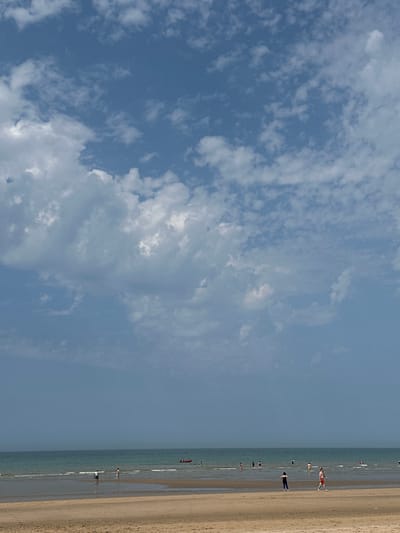 This image depicts a beach scene in Koksijde, Belgium. The foreground features a sandy beach with subtle ripples from receding waves. In the mid-ground, the sea is calm with gentle waves lapping the shore. Several people are visible in the shallow water and on the sand, some appearing to be wading or walking. A small red boat is present on the water in the distance, along with other distant figures. The sky above is a clear blue, dotted with scattered white clouds. The overall atmosphere is tranquil and suggestive of a pleasant day at the seaside.