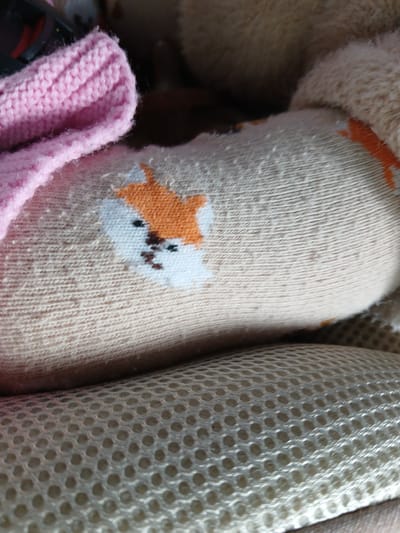 This is a close-up, macro photograph of a light brown sock with a small, embroidered orange and white fox face. The sock is slightly bunched and appears to be made of a textured knit material. A pink, ribbed knitted cuff is visible on the left side of the image, partially overlapping the sock. Below the sock, a beige, mesh-like material with a hexagonal pattern is visible. The background is dark and out of focus, suggesting an indoor setting. There are no people, actions, or interactions depicted. The location of Sorochkino, Russia is not evident from the visual information in the image.