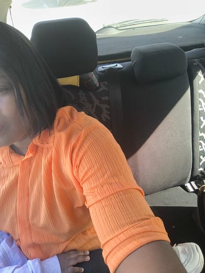 The image captures the interior of a vehicle, specifically the rear passenger area, in the city of Kızılbaş, Northern Cyprus / Cyprus. A female individual, partially depicted on the left, wears an orange, textured, long-sleeved shirt. Her dark, straight hair is visible at her shoulder. A child, whose left arm, hand, and a foot wearing a white sneaker are visible, is positioned near the adult's lap. The child wears a light blue or purple long-sleeved garment. The vehicle's interior features dark grey fabric seats with a subtle patterned section. Two dark grey headrests are visible, and a black seatbelt crosses the seat. Direct sunlight enters from the right, casting a distinct diagonal shadow across the right portion of the rear seat. A small, dark object with red trim is present on the seatback behind the adult. A dark bag with a light-colored strap is partially visible on the far right. The exterior through the rear window appears brightly lit and out of focus.