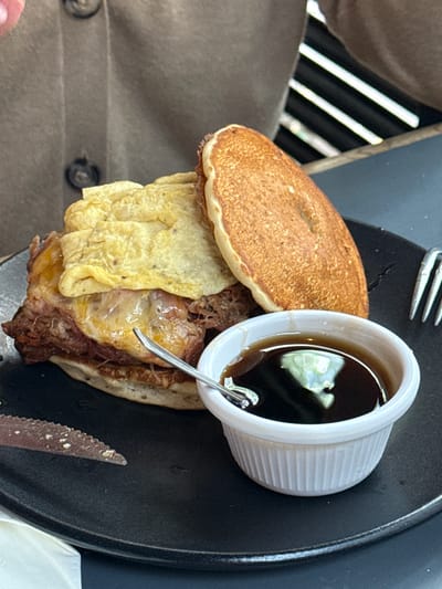 The image depicts a food item, seemingly a sandwich of sorts, served on a black plate. The sandwich consists of two pancake-like discs with a filling of shredded meat and a yellow, egg-like component. A small white ramekin filled with a dark liquid, possibly a dipping sauce or syrup, sits on top of the sandwich. A silver fork is visible at the top of the plate, and a knife is located at the bottom right. The setting appears to be an outdoor dining area, with a person wearing a brown jacket seated nearby. The image does not provide enough information to confirm that the location is La Tijera, Mexico.
