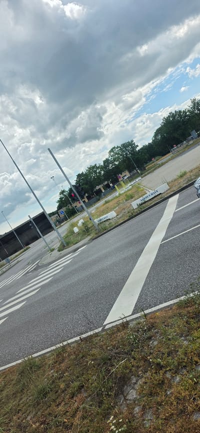 The image shows a street scene in Aarhus, Denmark. The perspective is from a high angle, looking down at the street, which is paved and has a white diagonal marking across it, likely indicating a pedestrian crossing. On the left side of the street, there are trees and some structures, possibly buildings or shelters. There are also traffic lights visible, one of which is red. Several tall metal poles are present, running parallel to the road. Along the edge of the road, there is dry grass and some small white flowers. The sky is cloudy, with patches of blue visible, suggesting a partly cloudy day.
On the right side of the road, there are white rectangular signs with text. One sign clearly reads "AARHUS HAVN" multiple times. This indicates the location is near the Port of Aarhus. There are no people clearly visible in the image. The scene appears to be an urban or semi-urban environment, possibly on the outskirts of the city or near a commercial/industrial area connected to the port. The lighting suggests it is daytime, but the cloudy conditions might make it difficult to pinpoint the exact time. The overall mood of the image is neutral, focusing on the details of the street and its surroundings.