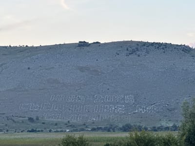 The image depicts a hillside in Aldomirovtsi, Bulgaria, with large white lettering carved into the rocky surface. The lettering appears to be a memorial or celebration, displaying the dates "1885-2025" on the top line and "СЪЕДИНЕНИЕ НА БЪЛГАРИЯ" (which translates to "Unification of Bulgaria") on the lines below. The hillside itself is covered in stones and sparse vegetation, with a few scattered trees and bushes visible on the lower slopes. In the foreground, there is a field of grass and more trees. The sky is overcast with a pale, hazy light.