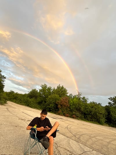 The image displays an outdoor scene featuring a young man seated on a portable chair under a prominent double rainbow, likely in Tuchenitsa, Bulgaria. The man, dressed in a black t-shirt, black shorts, and black athletic shoes, is absorbed in his smartphone, which he holds in both hands. A bottle of a green-yellow sports drink with an orange cap (resembling Gatorade) is visible, hanging from the side of his chair.

He is positioned on a cracked, grey asphalt or concrete surface, which shows faint, dark, circular tire-like marks. Lush green trees, some with reddish-brown foliage, border this open area, providing a natural backdrop.

The sky above features a striking double rainbow, arching from the upper left to the lower right of the frame. The clouds are a mix of light grey and soft, warm-toned patches, suggesting either clearing weather after rain or the soft light of late afternoon or early evening. The presence of the rainbow strongly indicates recent precipitation. In the far upper left corner of the sky, a very faint and indistinct object, possibly a distant bird or aircraft, can be seen. The overall impression is one of peaceful observation, with the natural spectacle of the rainbow providing a serene atmosphere.