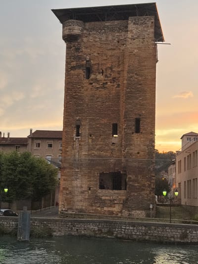 The image shows a tall, stone tower in Vienne, France, at dusk. The tower is made of rough, reddish-brown stone blocks and has a dark, sloping roof. There are several small, rectangular openings, or windows, on the face of the tower. At the top left of the tower, there is a small, rounded turret-like structure. In front of the tower, there is a body of water, likely a river, with a stone wall bordering it. Beyond the tower, several buildings are visible, including a row of houses with red tile roofs and a larger, modern-looking building. Two green streetlights are visible on the right side of the image. The sky is a soft orange and grey, indicating the setting sun. The scene appears peaceful and historic.